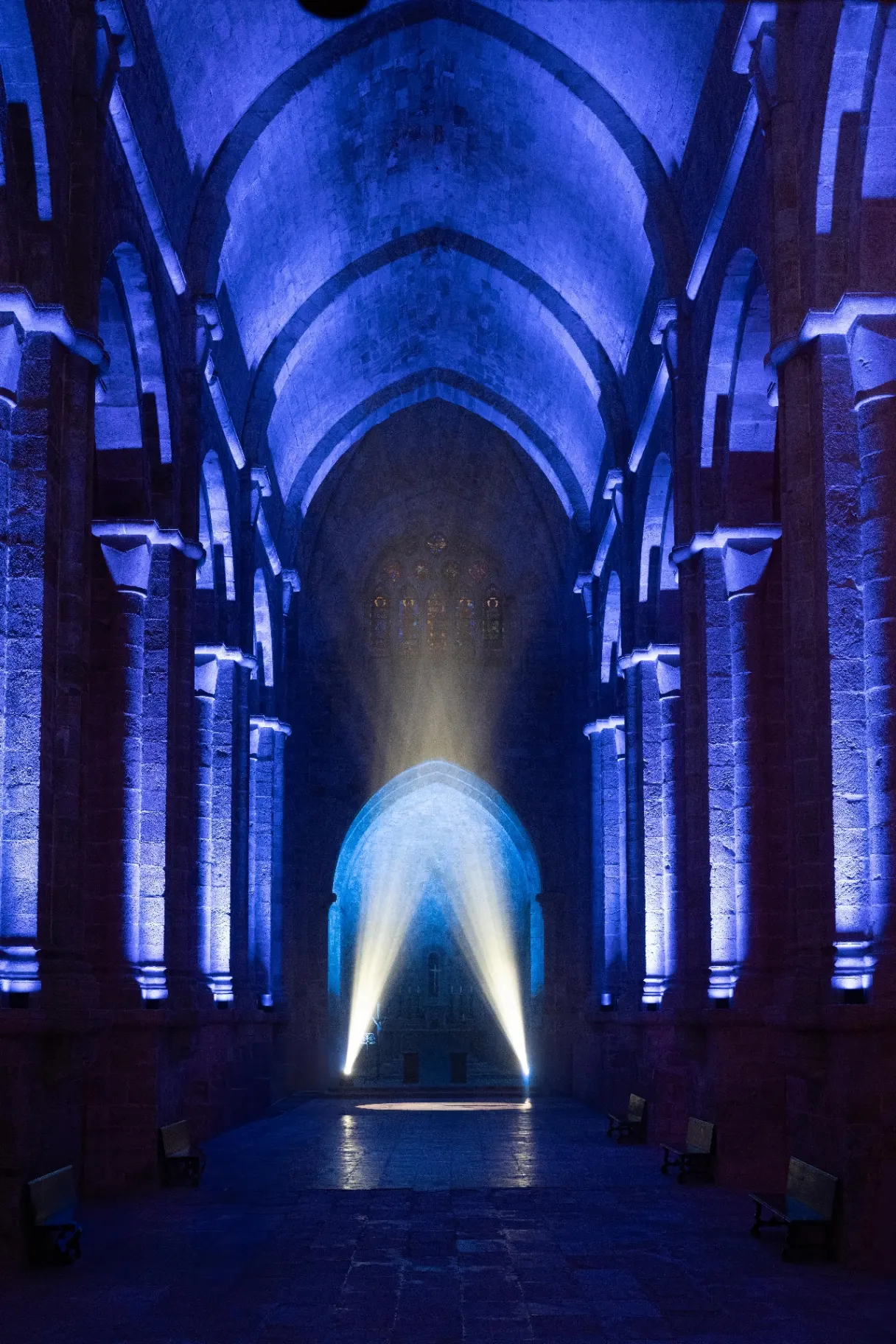 Nef gothique éclairée en bleu avec deux faisceaux lumineux blancs convergeant vers le sol, Abbaye de Fontfroide.